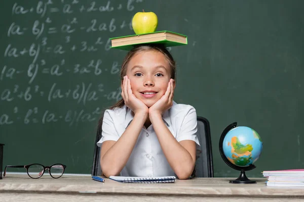 Scolaretta Giocosa Seduta Alla Scrivania Con Libro Mela Testa — Foto Stock