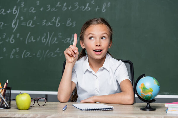 amazed schoolgirl showing idea gesture near globe, notebook and blurred chalkboard