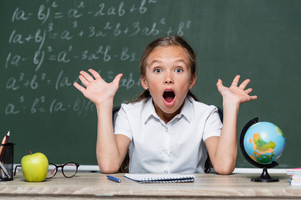 amazed schoolgirl with open mouth gesturing while sitting at desk in classroom