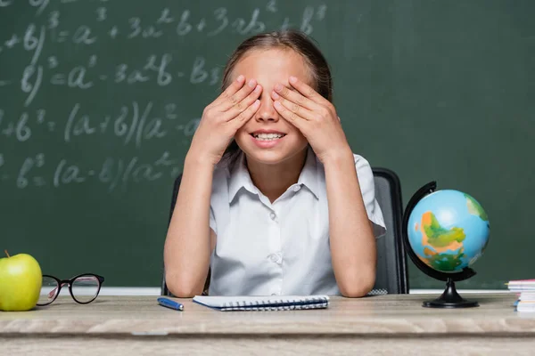 Cheerful Schoolgirl Covering Eyes Hands Globe Notebook Desk — Foto de Stock