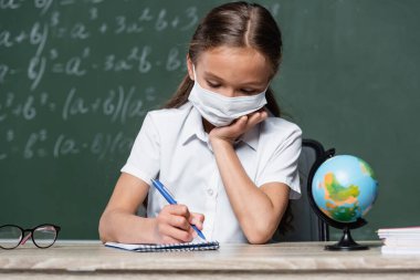 schoolchild in medical mask writing in notebook near globe and chalkboard on blurred background clipart