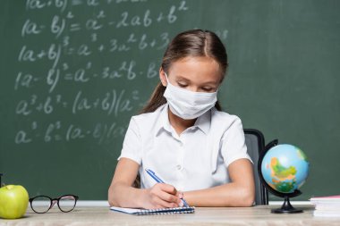 Tıp maskesine bürünmüş genç bir kız not defterine dünya, elma ve bulanık karatahta gibi şeyler yazıyor.