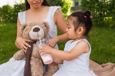 Asyalı küçük kız elinde plastik bardakla oyuncak ayının yanında annesinin ellerinde.