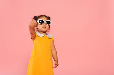 Sarı elbiseli ve güneş gözlüklü Asyalı çocuk pembe üzerinde baş parmağını gösteriyor.