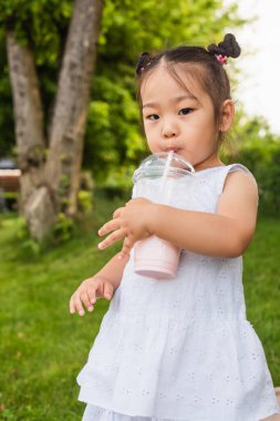 Asyalı çocuk elbise giymiş parkta samandan tatlı milkshake içiyor.