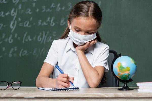 schoolchild in medical mask writing in notebook near globe and chalkboard on blurred background