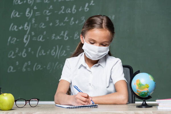 preteen schoolgirl in medical mask writing in notebook near globe, apple and blurred chalkboard