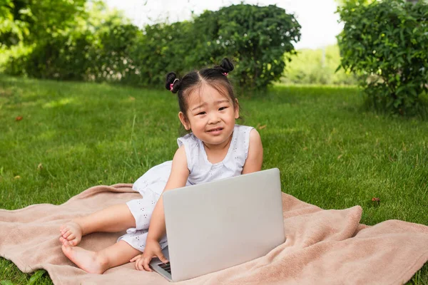 confused asian kid looking at camera near laptop on picnic blanket