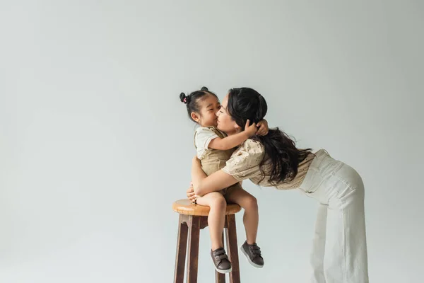 アジア系の幼児の女の子は椅子に座ってグレーで隔離された母にキス — ストック写真