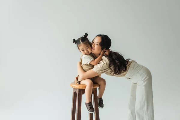 Felice Asiatico Bambino Ragazza Seduta Sedia Abbracciando Madre Isolato Grigio — Foto Stock