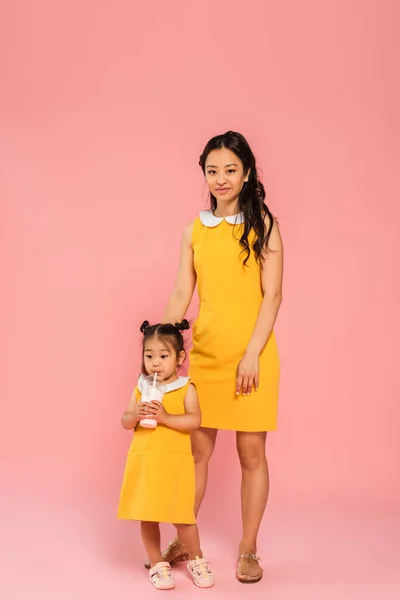 亚洲蹒跚学步的孩子穿着黄色衣服 用稻草喝着美味的奶昔 幸福的母亲在粉红上 — 图库照片