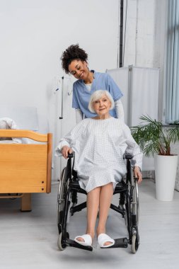 Klinikte tekerlekli sandalyedeki yaşlı hastanın yanında gülümseyen Afrikalı Amerikalı hemşire. 