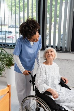 Afrika kökenli Amerikalı hemşire önlüklü ve tekerlekli sandalyedeki yaşlı hastaya bakıyor. 