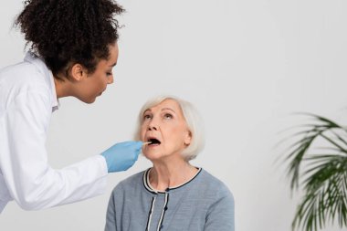 Lateks eldivenli Afrikalı Amerikalı doktor yaşlı hastanın boğazını kontrol ediyor.  