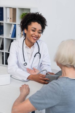 Gülümseyen Afrikalı Amerikalı doktor klinikte bulanık yaşlı kadına tonometre takıyor. 