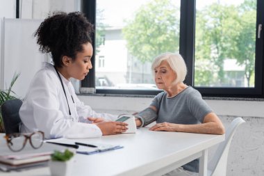 Afrikalı Amerikalı doktor yaşlı kadının tonometredeki basıncını kontrol ediyor. 