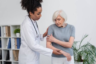 Senior patient touching arm near african american traumatologist in clinic  clipart