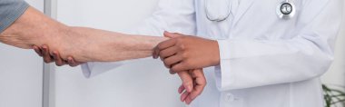 Cropped view of african american doctor holding injured hand of senior patient, banner  clipart