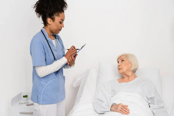 非洲裔美国护士在病床上靠近老年病人的写字板上写字 — 图库照片
