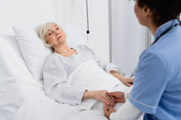 Senior patient holding hand of african american nurse in hospital ward 
