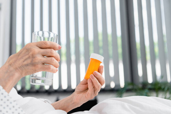 Cropped view of senior woman holding water and pills in hospital ward 