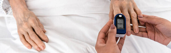Cropped view of african american nurse fastening digital oximeter on finger of woman in clinic, banner 