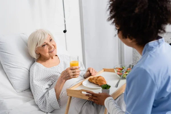 Paciente Anciano Sonriente Con Vaso Jugo Naranja Mirando Borrosa Enfermera — Foto de Stock