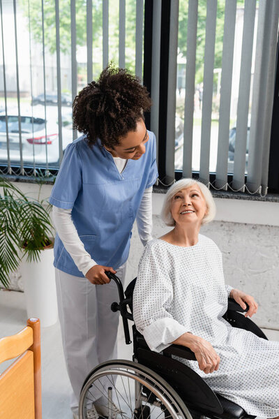 African american nurse looking at elderly patient in gown and wheelchair 