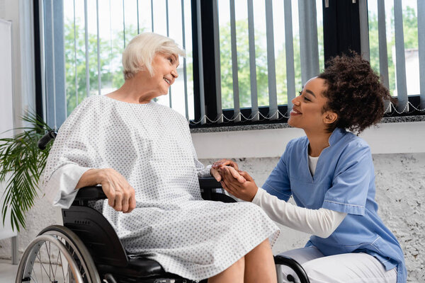 African american nurse holding hand of smiling elderly woman in wheelchair 