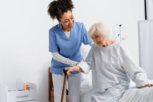 Smiling african american nurse helping patient with walking cane near hospital bed 