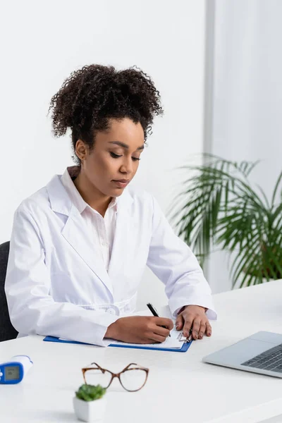 Médico Afroamericano Bata Blanca Escribiendo Portapapeles Cerca Laptop Pirómetro — Foto de Stock