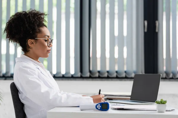 非洲裔美国医生拿着笔靠近笔记本电脑和模糊的高温计的侧视图 — 图库照片