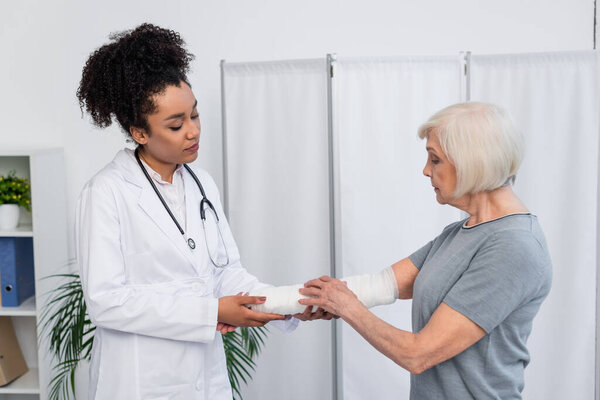 African american traumatologist looking at plaster bandage on arm of senior patient 