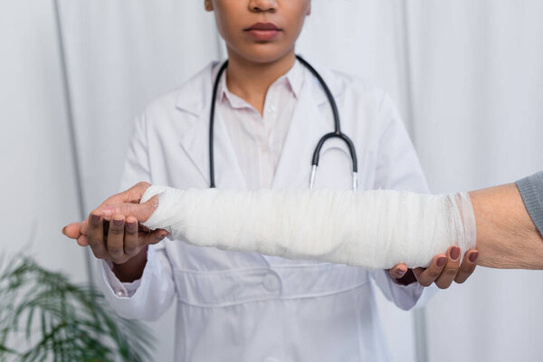 Cropped view of blurred african american doctor touching hand of elderly woman in plaster bandage 