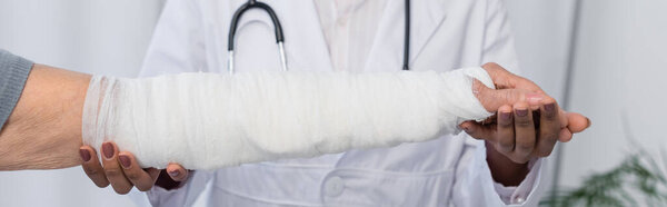 Cropped view of african american traumatologist holding hand of patient in plaster bandage, banner 