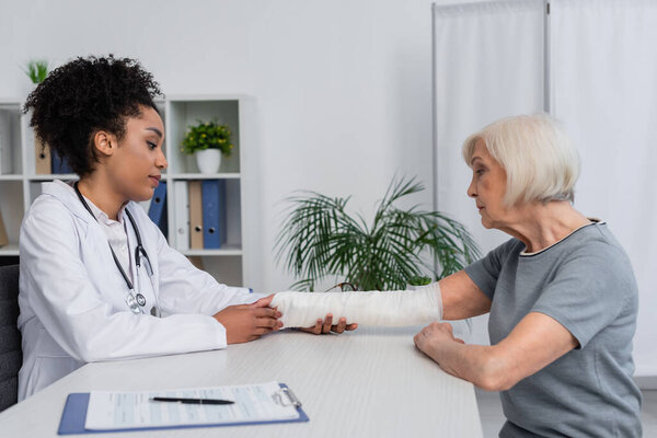 Side view of african american traumatologist in white coat checking broken arm of senior woman 