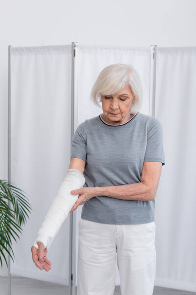 Elderly woman touching arm in plaster bandage in hospital 