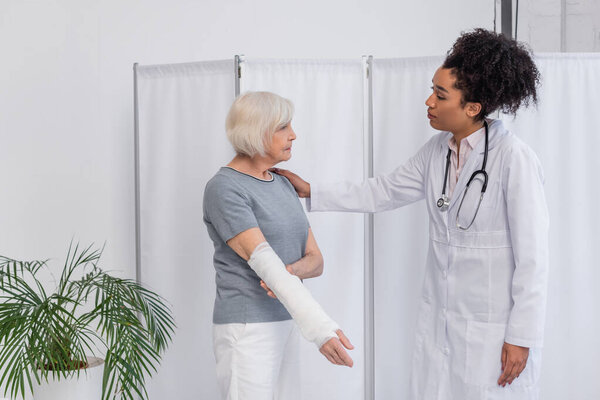 Side view of african american doctor calming patient with plaster bandage on arm in clinic 