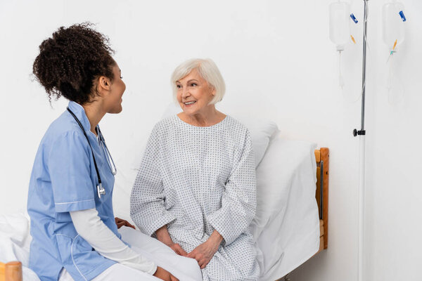 Senior patient looking at african american nurse on hospital bed 