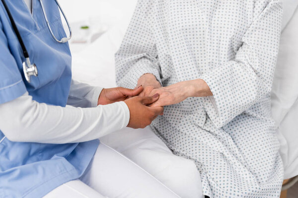 Обрезанный вид африканской американской медсестры, держащей за руки пожилого пациента в халате 