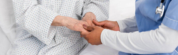 Обрезанный вид африканской американской медсестры, держащей за руки пациента в халате на кровати, баннер 