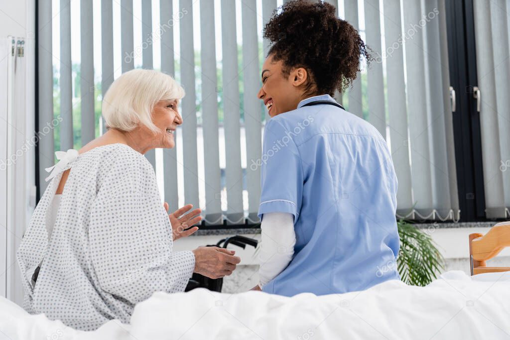 Side view of cheerful senior woman talking to african american nurse on bed 