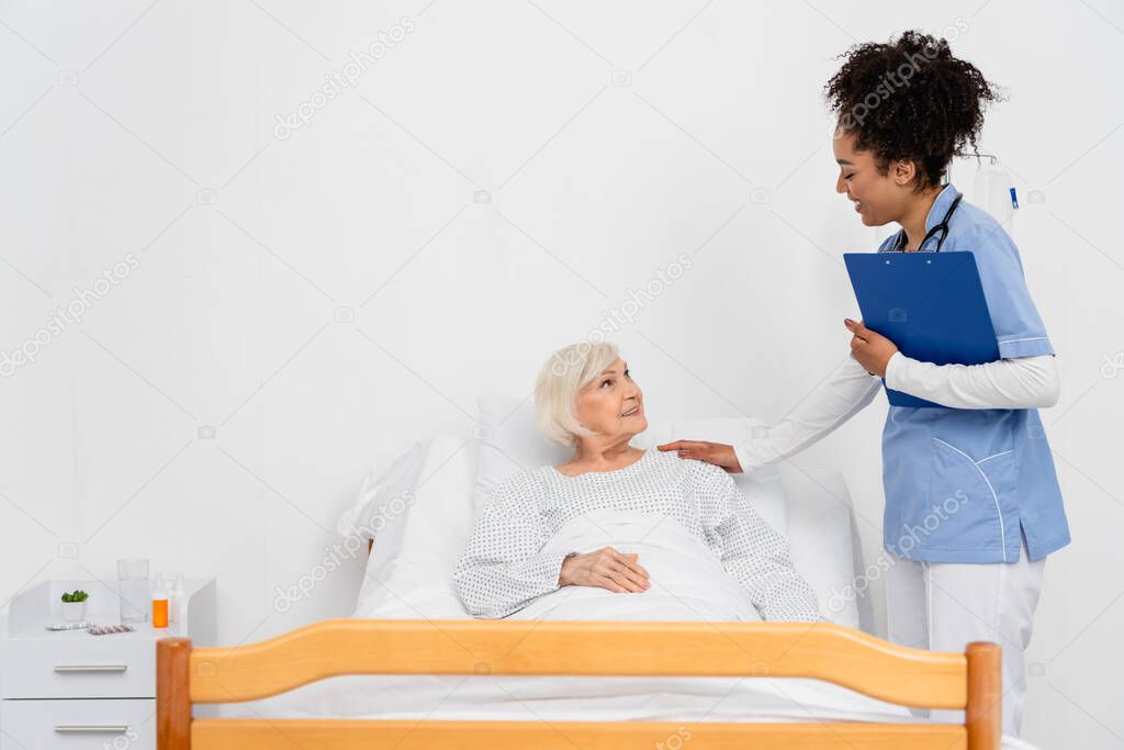 African american nurse with clipboard touching elderly patient on bed 