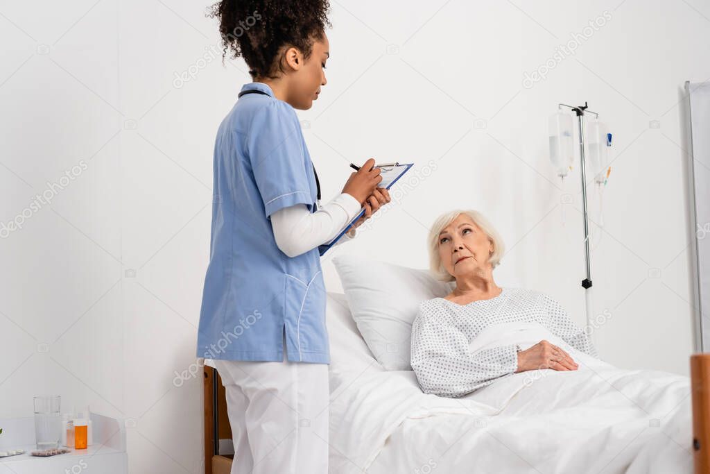 African american nurse writing on clipboard near elderly patient on hospital bed 