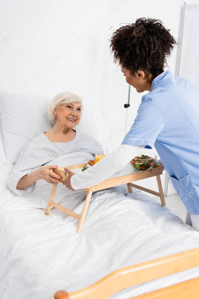 African american nurse holding tray with food near cheerful patient on bed in hospital ward 