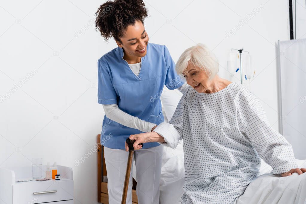 Smiling african american nurse helping patient with walking cane near hospital bed 