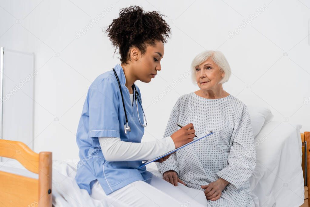 African american nurse writing on clipboard near elderly woman in hospital bed 