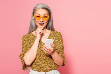 Pembe üzerine izole akıllı telefon kullanarak çeneye dokunan neşeli Asyalı kadın.