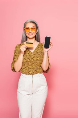 Kredi kartı ve akıllı telefonu olan olgun Asyalı bir kadın pembe arka planda kameraya gülümsüyor.
