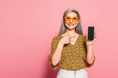 Şık güneş gözlüklü gülümseyen Asyalı kadın pembe arka plandaki akıllı telefonu işaret ediyor.